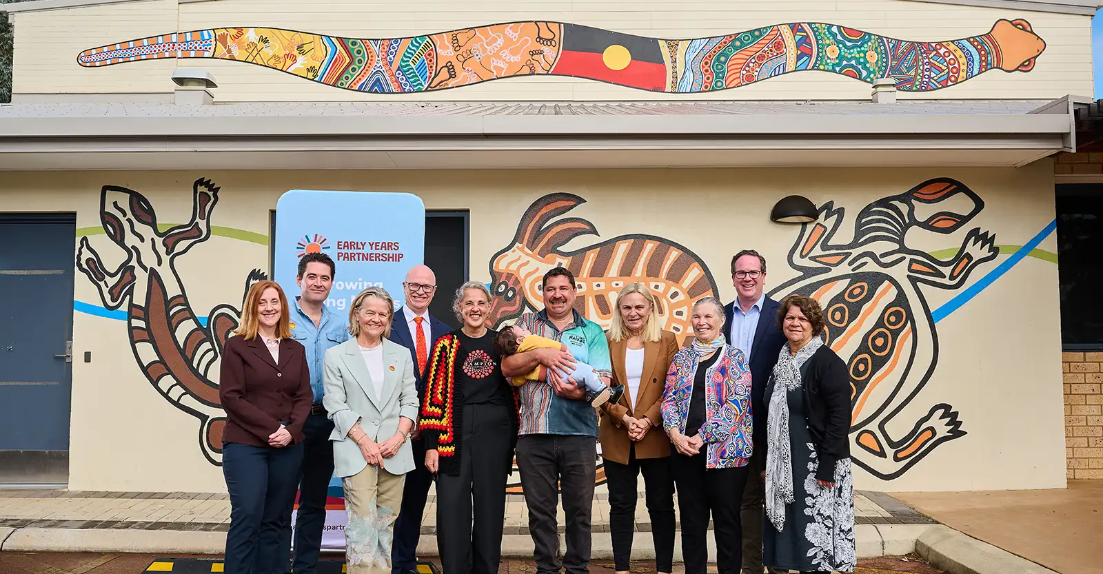 A group of men and women standing outside a community centre that is painted with animals in the First Nations style, including the Waygl or Rainbow Serpent, with the Australian Indigenous flag as part of the pattern on its body.