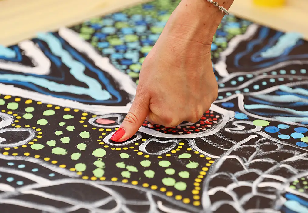 A close up of a woman's hand, pressing her thumb onto a canvas painted with a First Nations design, to add to the painting.