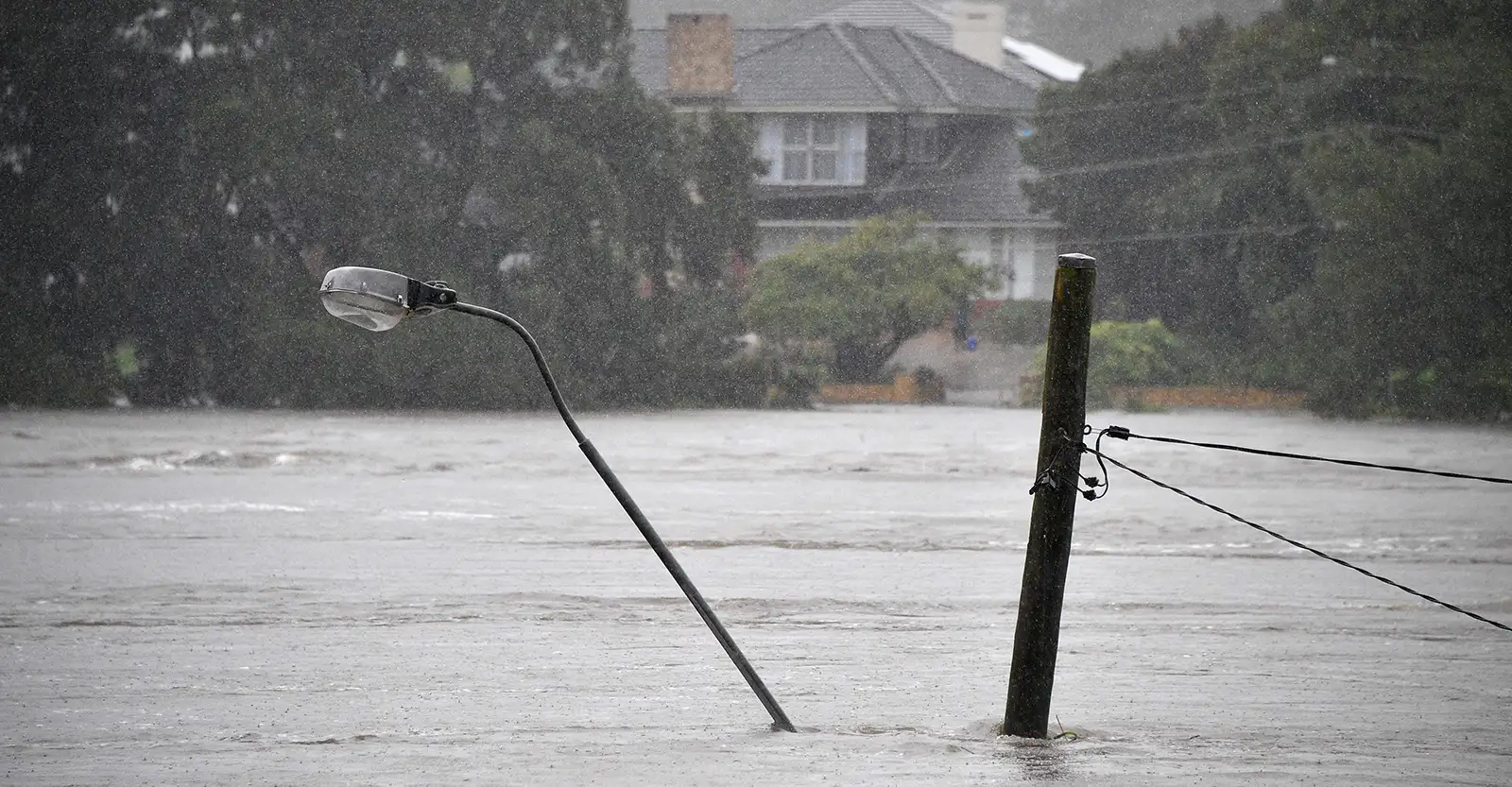 A photo of a light pole sticking out of very high level flood waters. In the background is a two storey house on slightly higher ground, and it can be seen that it is still raining.