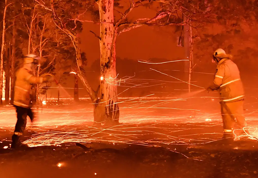 Two fire fighters spray water on a bushfire. The photo has a slow shutter speed so that bright sparks of fire have made glowing trails. The light is extremely orange, making the whole photo have a strong orange cast. The mood of the photo is of a scary and dangerous situation.