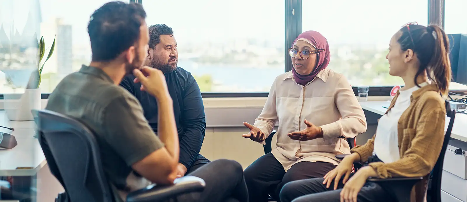 A small multiracial group of men and women sit in a circle on office chairs, turned around from their desks, having a conversation. A woman wearing a hijab is talking and gesturing with her hands.