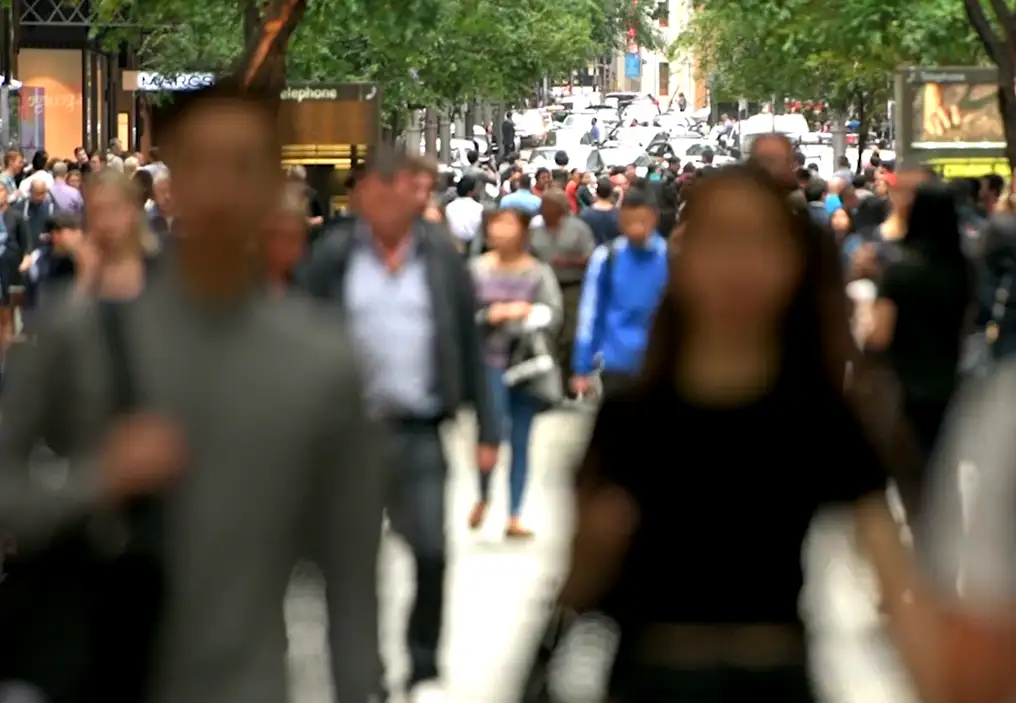 A crowd of people walking along a pedestrian mall. The plane of focus is on the leafy trees in the background, so all the people's faces are blurred.