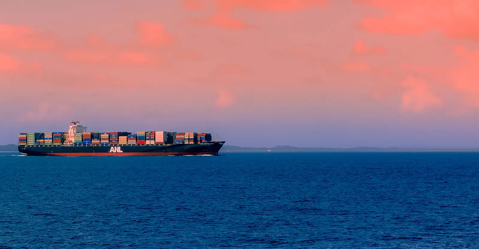 A long shot of a large cargo ship loaded with shipping containers on a calm ocean at sunset. The ocean is dark blue and the sky is a rosy pink and purple.