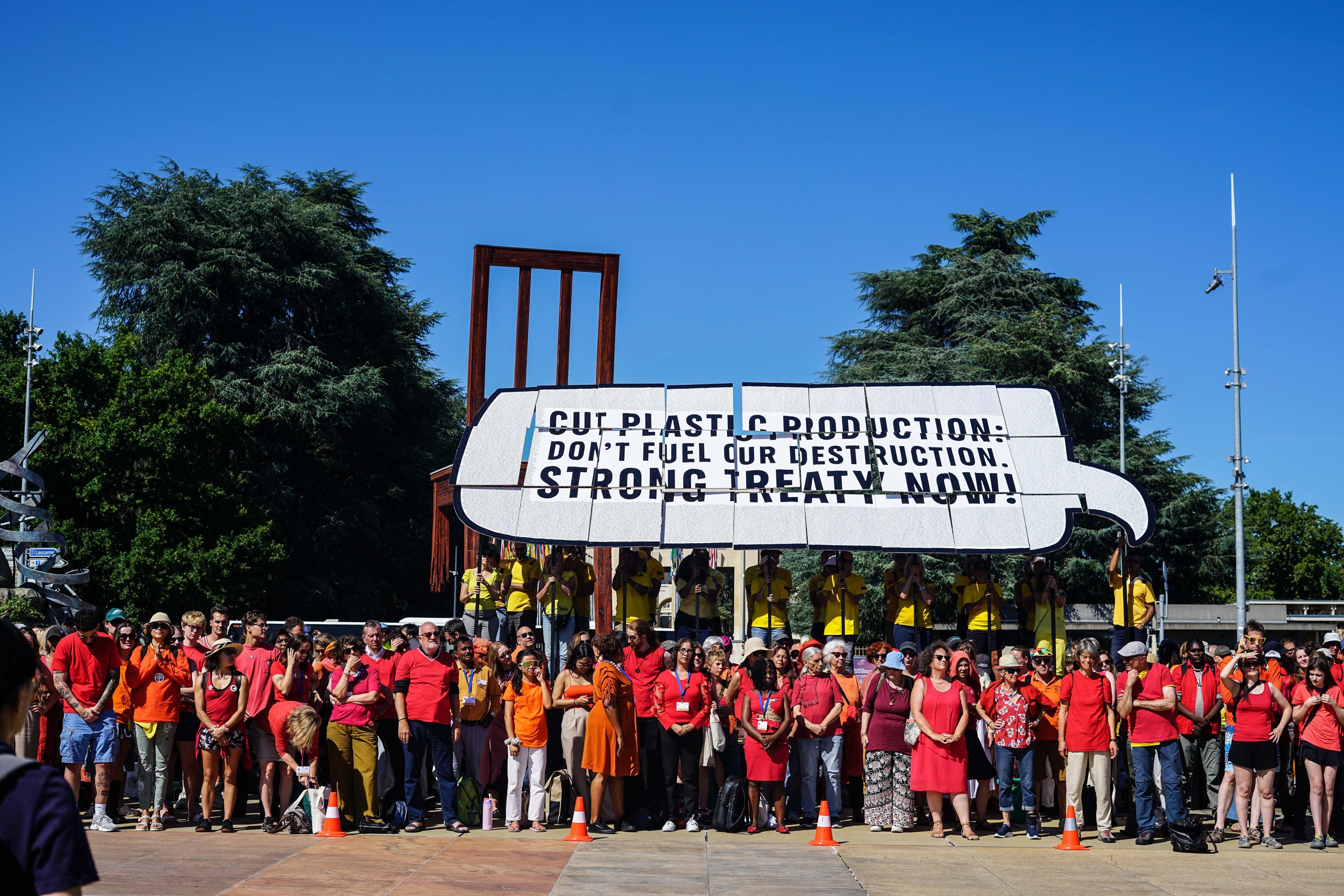 A large group of activists wearing bright red and orange stand in front of another group of people wearing yellow t-shirts and each holding a pole with a card attached to it. Held together, the cards depict a speech bubble that says “Cut plastic production: don't fuel our destruction. Strong treaty now!” The speech bubble appears to come from the protester to the very right of frame, who is holding the card with the tail of the speech bubble.
