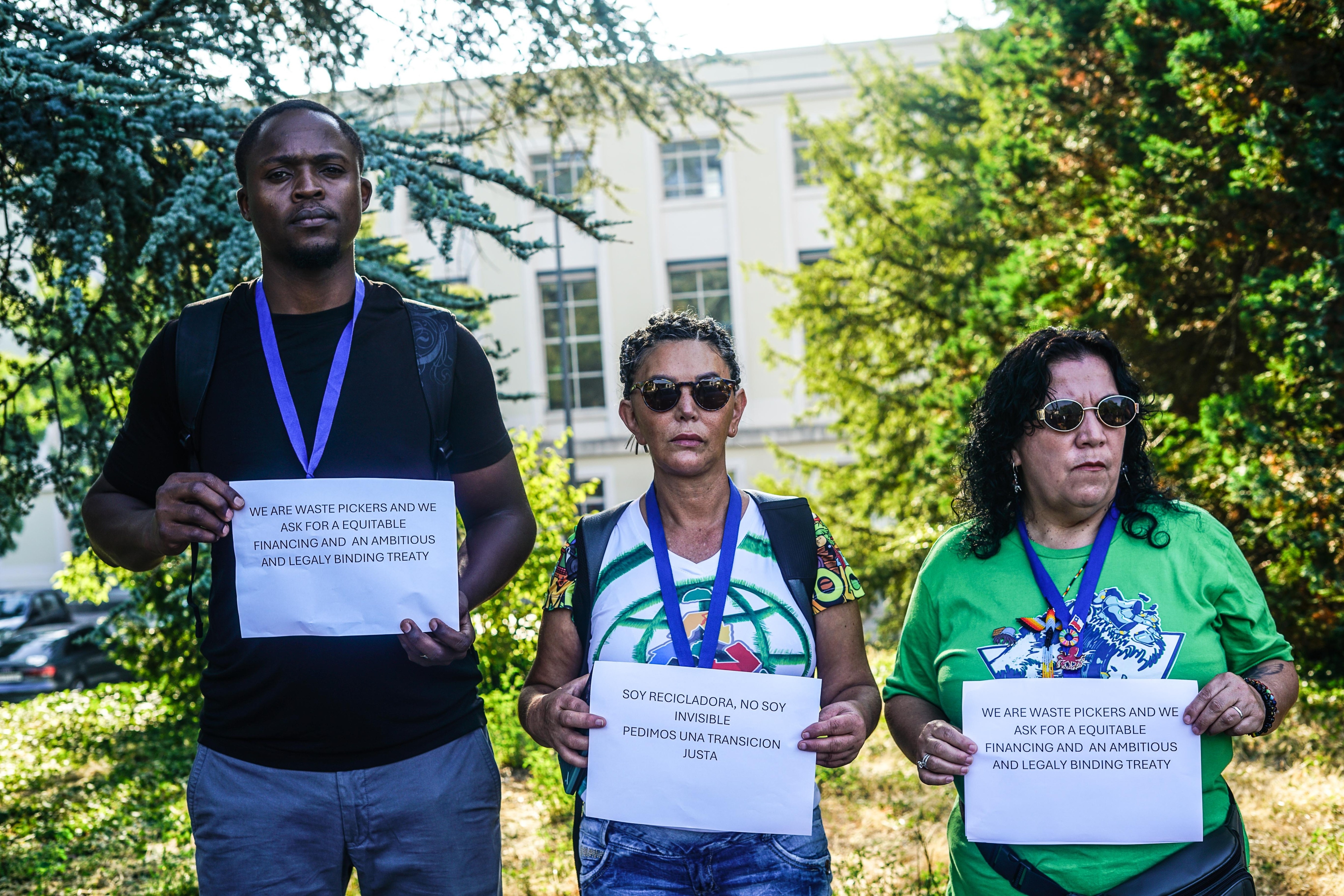 Three people stand outside the UN building in Geneva holding A4 pieces of paper that say “We are waste pickers and we ask for a equitable financing and an ambitions and legaly binding treaty [sic]” and “Soy recicladora, no soy invisible. Pedimos una transicion justa” (“I am a waste picker, I am not invisible. We ask for a just transition"). They look stern and determined.