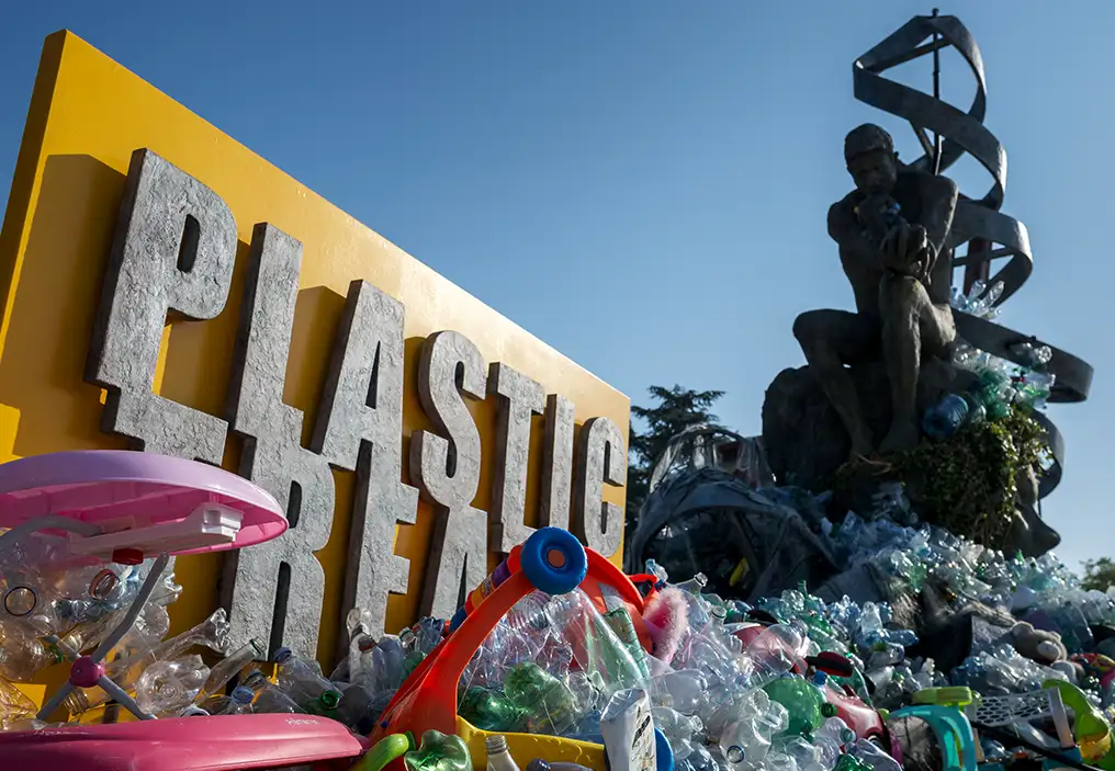 A low angle shot of Ben Von Wong’s installation entitled The Thinker’s Burden. It is a 6m tall facsimile of Rodin’s The Thinker, with winding flat strips of bronze representing a DNA double helix wrapped around the Thinker. At the foot of the sculpture and the base of the seat is plastic waste, mostly single-use drink bottles, which over the course of a week was added to, to completely eclipse the Thinker himself. Behind the installation is a large yellow board with the words PLASTIC TREATY on it.