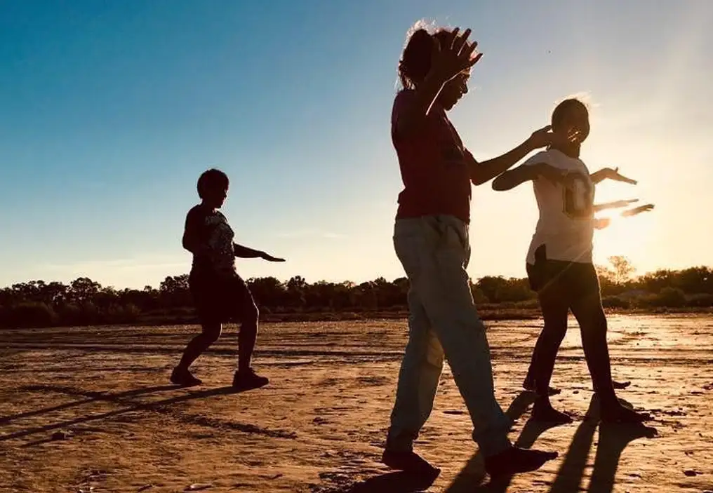 Four First Nations children perform on a clay pan in a remote site in New South Wales, at sunset. They are walking with their hands held out in front of them, parallel to the ground. The sun is low in the sky and creates a warm glow over the image, and means the children are back lit.