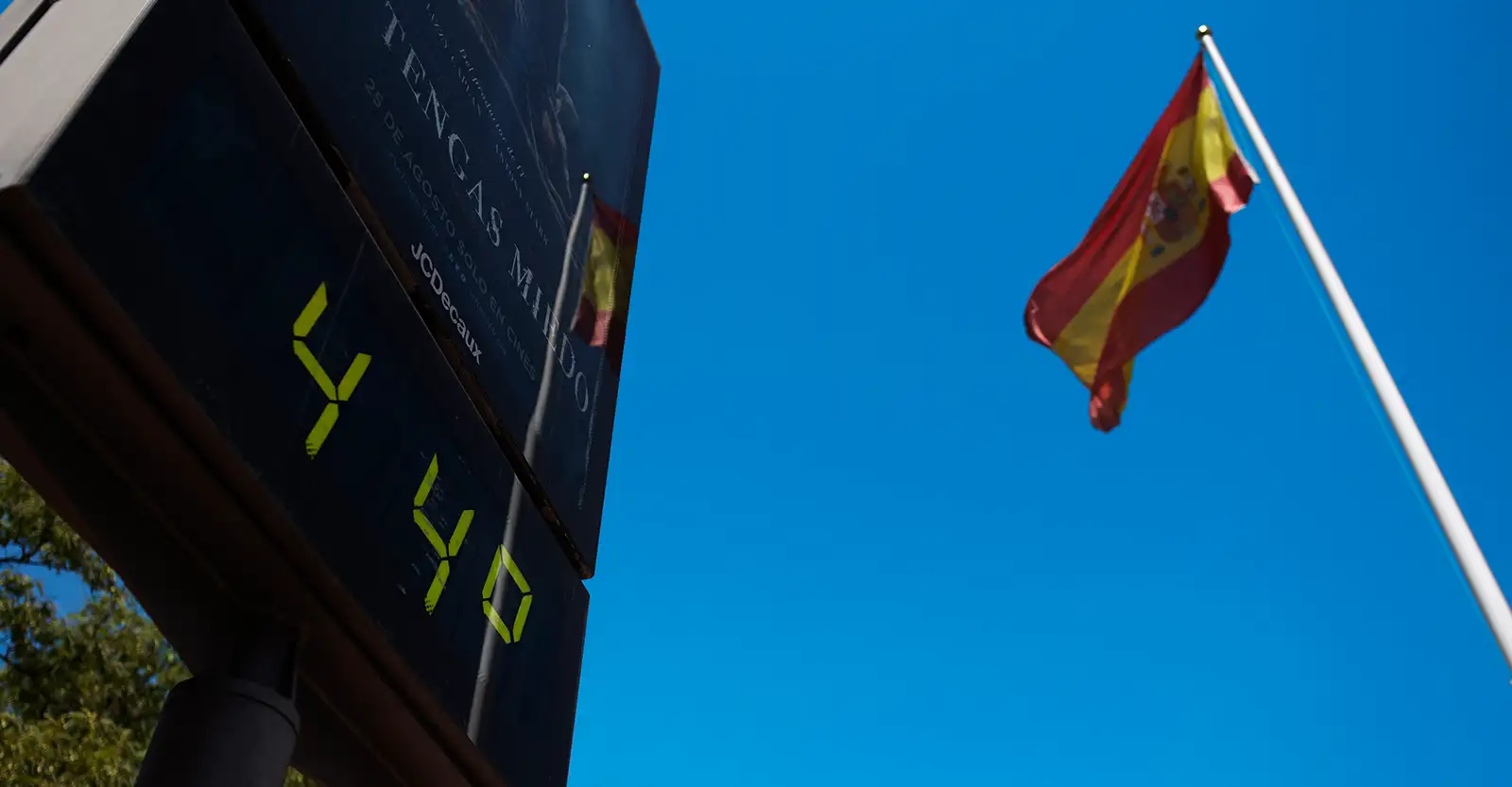 A low angle shot of a digital board showing that the temperature is 44 degrees Celcius. Next to the board is a flag pole flying the red and yellow flag of Spain.