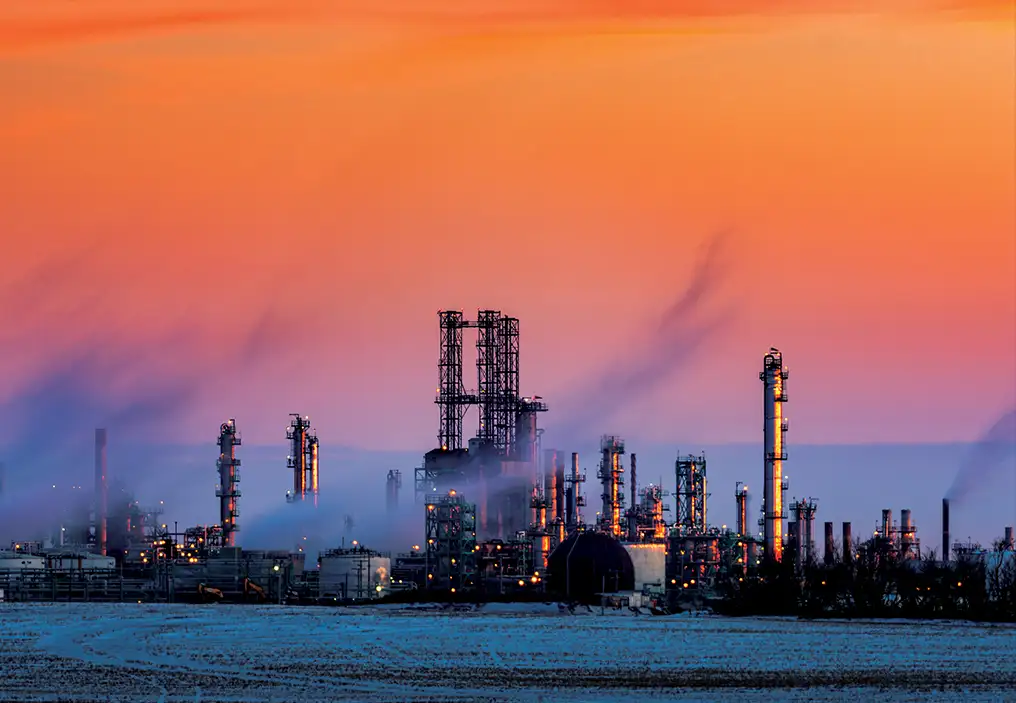 A refinery complex at sunset. Across a field with frost on it, the industrial complex can be seen, putting out large amounts of heavy vapour. The sunset is beautiful and dramatic colours of pink and orange.