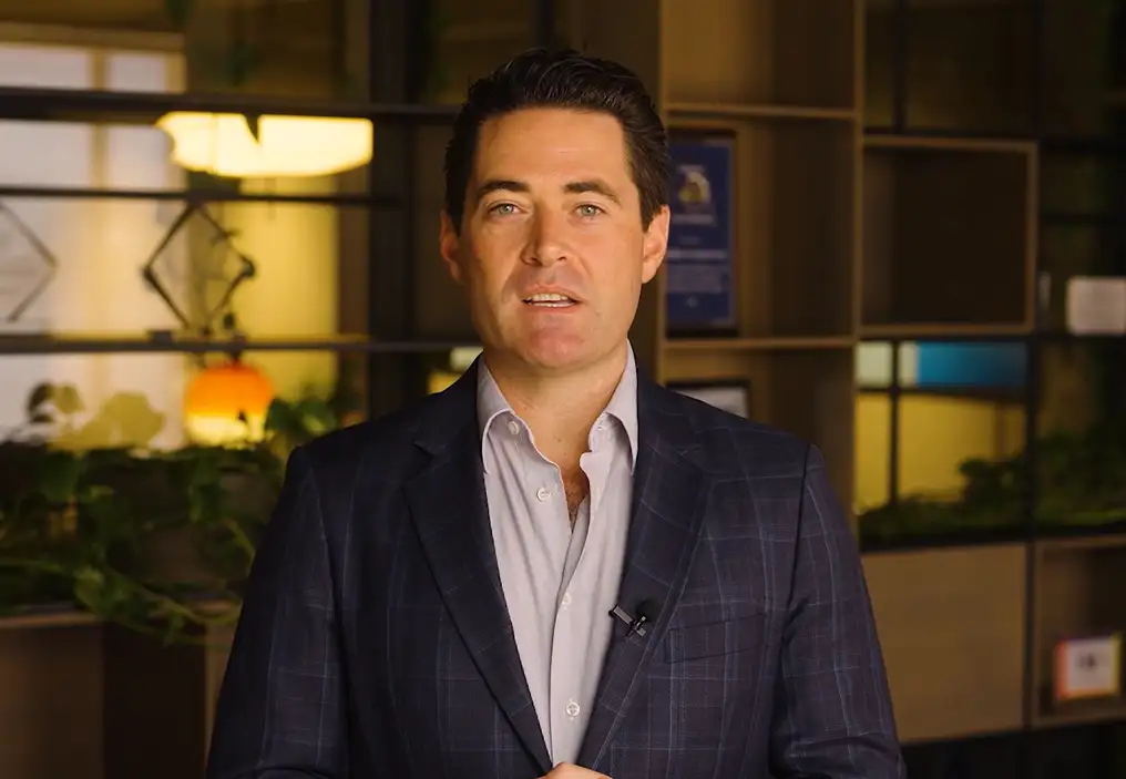 John Hartman looks at the camera, in the middle of speaking. He is wearing a dark blue checked sportscoat and a light blue shirt unbuttoned at the top. He looks professional but approachable.