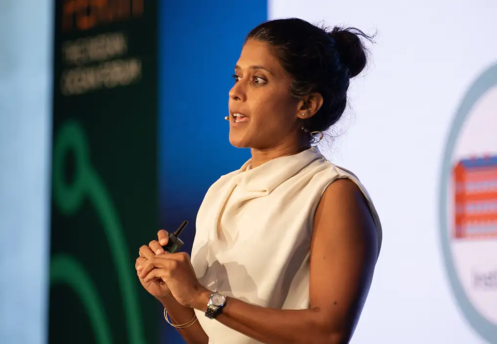 Dr Asha de Vos, a Sri Lankan woman, is speaking into a headset microphone, holding a clicker for a screen. The shallow depth of field means that the background is out of focus, but it can be seen she is standing in front of a slide deck and to the side is a range of pull up banners.