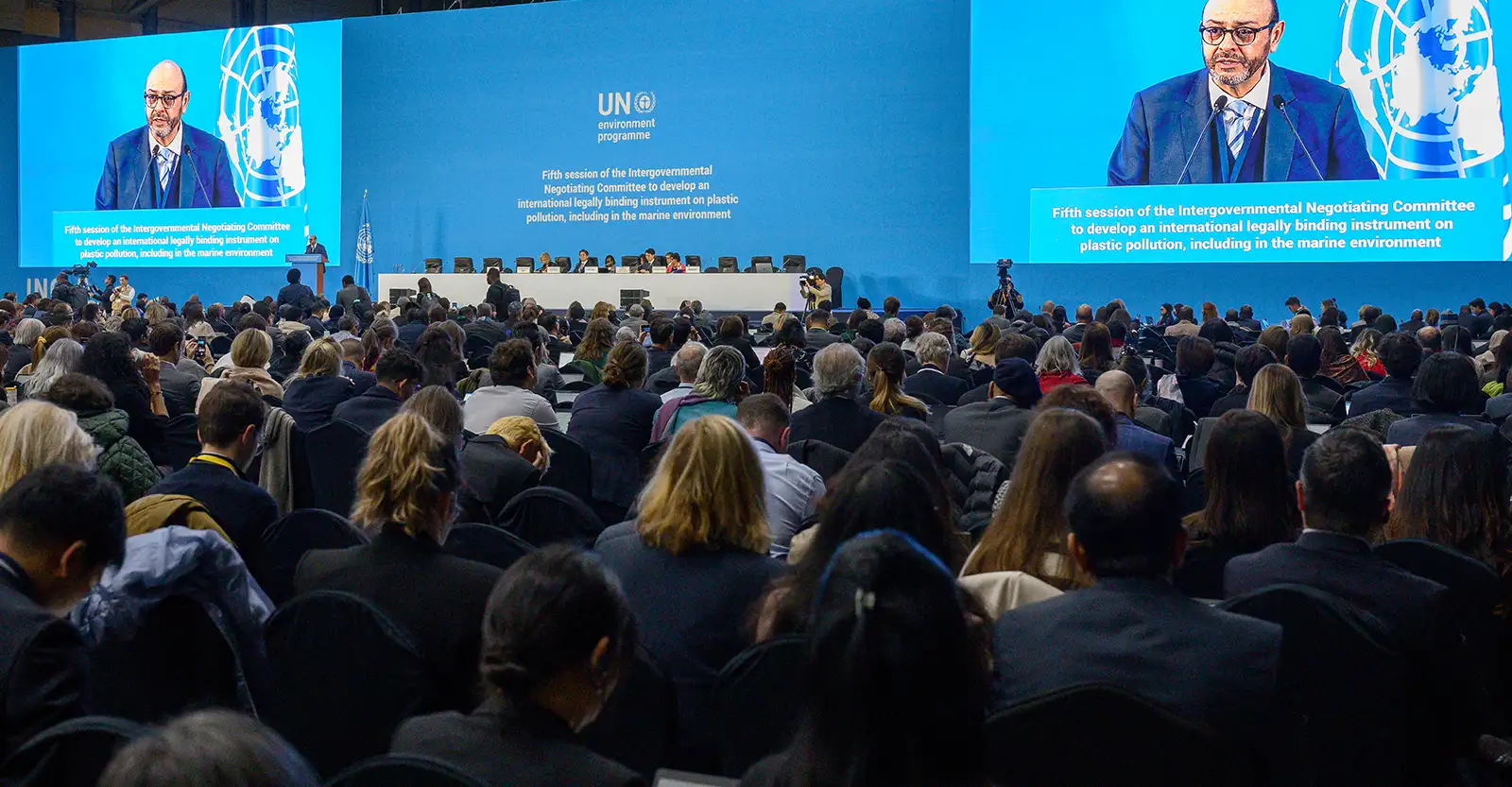 A photo taken from the back of an audience in a large conference room, looking towards a dais with a table with a group of panel speakers sitting at it. To the left of the table is a podium that Luis Vayas Valdivieso is speaking at. Behing the dais there is a large blue background with the full name of INC-5.1 on it, and to either side are large screens that allow the people to see a close up of Ambassador Valdivieso.