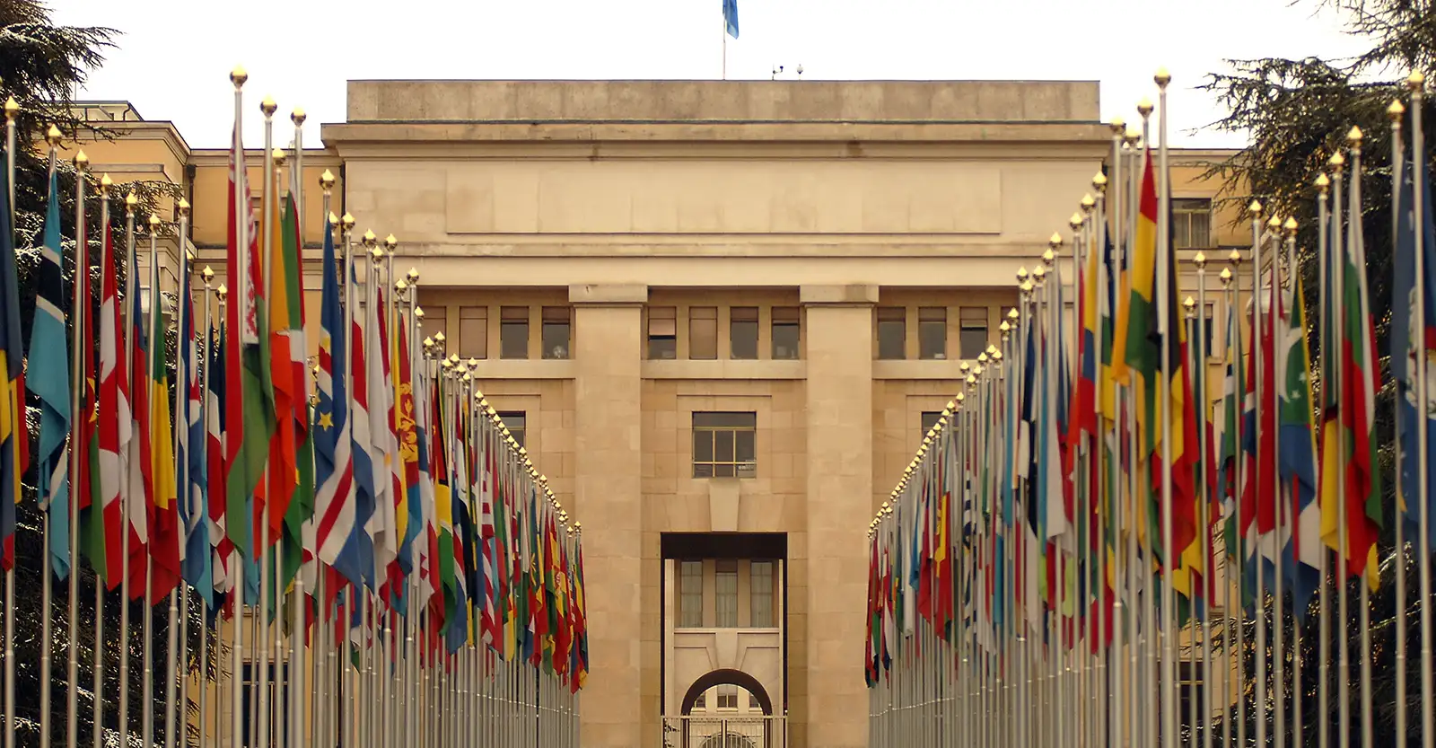 A view of the Palace of Nations, a stately sandstone building of linear architecture, from a promenade leading up to an entrance. The promenade has a double row of various nations' flags on either side of it, that are hanging stright downwards due to the lack of wind.