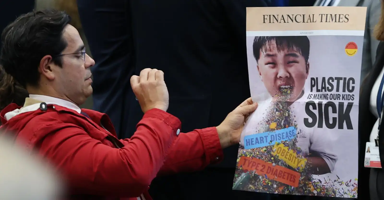 A side on shot of a man with black hair, wearing a bright red jacker. He is holding a wraparound of the Finanical Times in his left hand, and holding his phone up in his right to take a photo of it. The wraparound is a photo of a Korean boy, altered to make it seem as though he is vomiting small bit of plastic refuse. The headline reass 'Plastic is making our kids sick' and in the refuse the words 'Heart disease', 'Obsesity' and 'Type 2 diabetes', demonstrate just a few of the health effects that plastic can lead to.