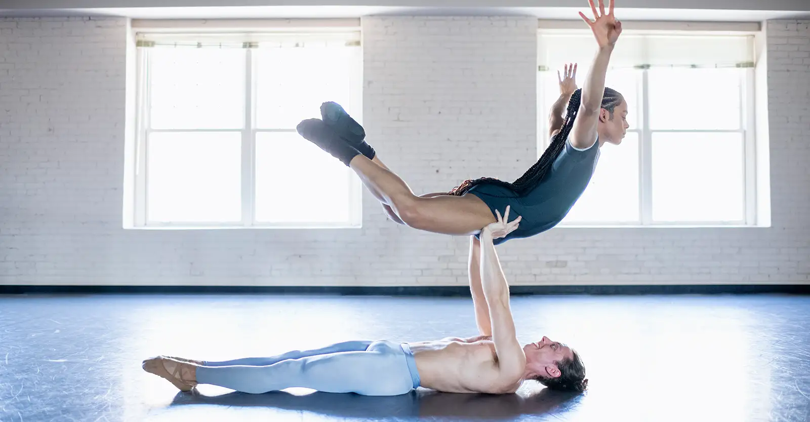 A male ballet dancer wearing only grey tights lies on his back on the floor of a ballet studio, lifting a non-binary ballet dancer with long dreadlocks who is wearing a dark grey leotard.