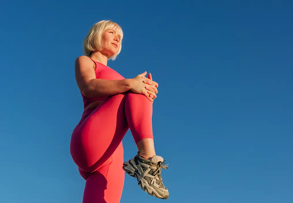 A woman wears a matching pink activewear set as she stretches her legs against a clear blue sky.