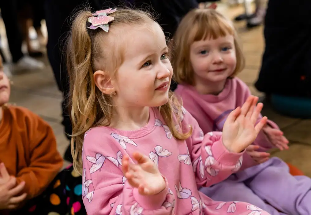 A little girl wearing a pink jumper looks up while pulling her hands apart to clap. In the background are another little girl and a little boy, also clapping. All three are sitting on the ground.