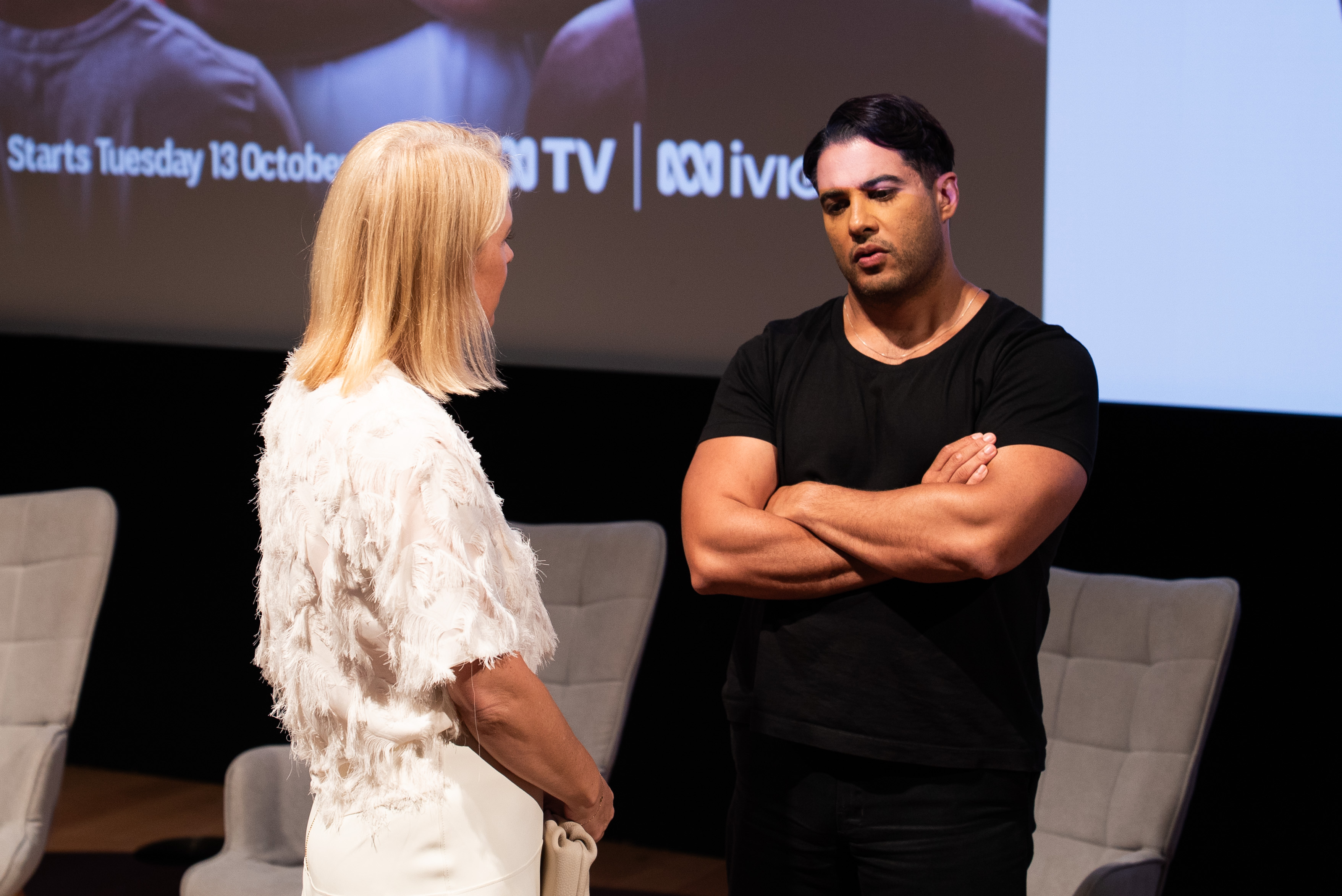 Marc Fennell stands with his arms crossed answering a question. In the background, you can see there is a Q&A panel before a movie theate screen with The State of Man branding.