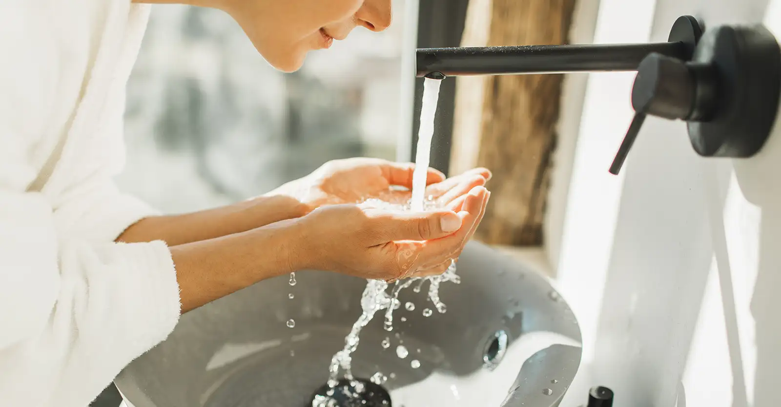 A woman leans over a grey round bathroom sink, cuppeing her hands under the water streaming out of the black tap. She is wearing a bathrobe and only the chin of her face can be seen.