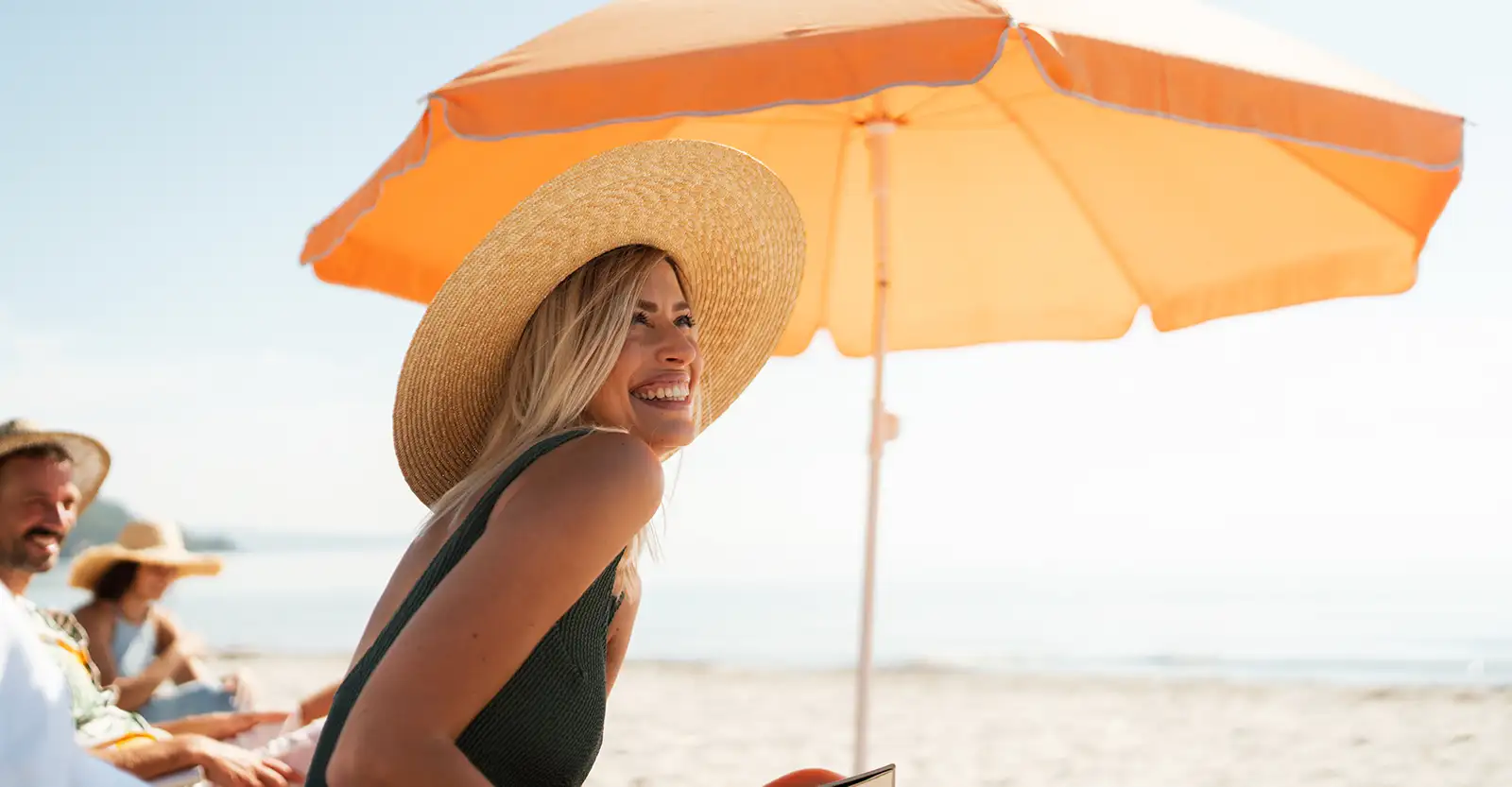 A blonde woman sits at the beach under a light orange umbrella and with a wide-brimmed straw hat for sun protection.