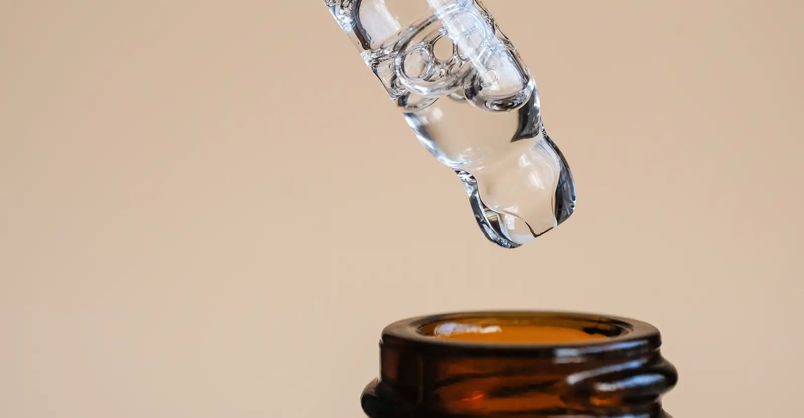 A close up of a glass eye-dropper with clear liquid in it, posed over the rim of a small brown glass bottle.