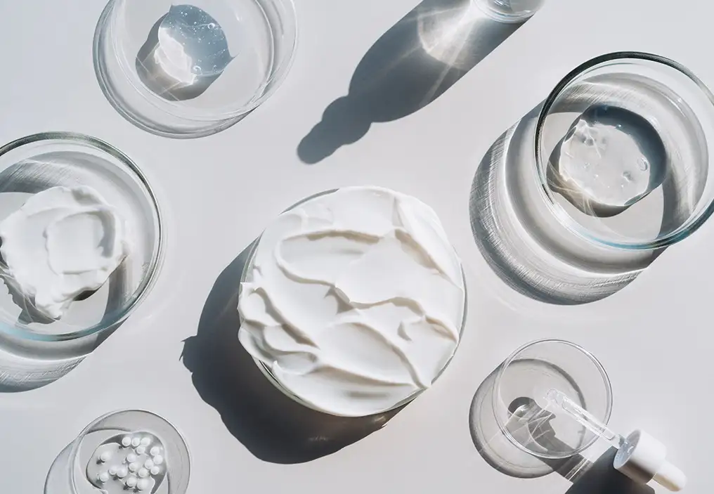 A bird's eye view of a range of creams, gels and liquids in petri dishes, on a white background. The effect is very scientific.