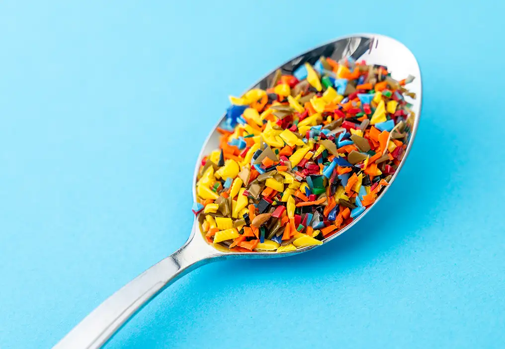 A metal spoon heaped with bright multi-coloured pieces of plastics on a light blue background.