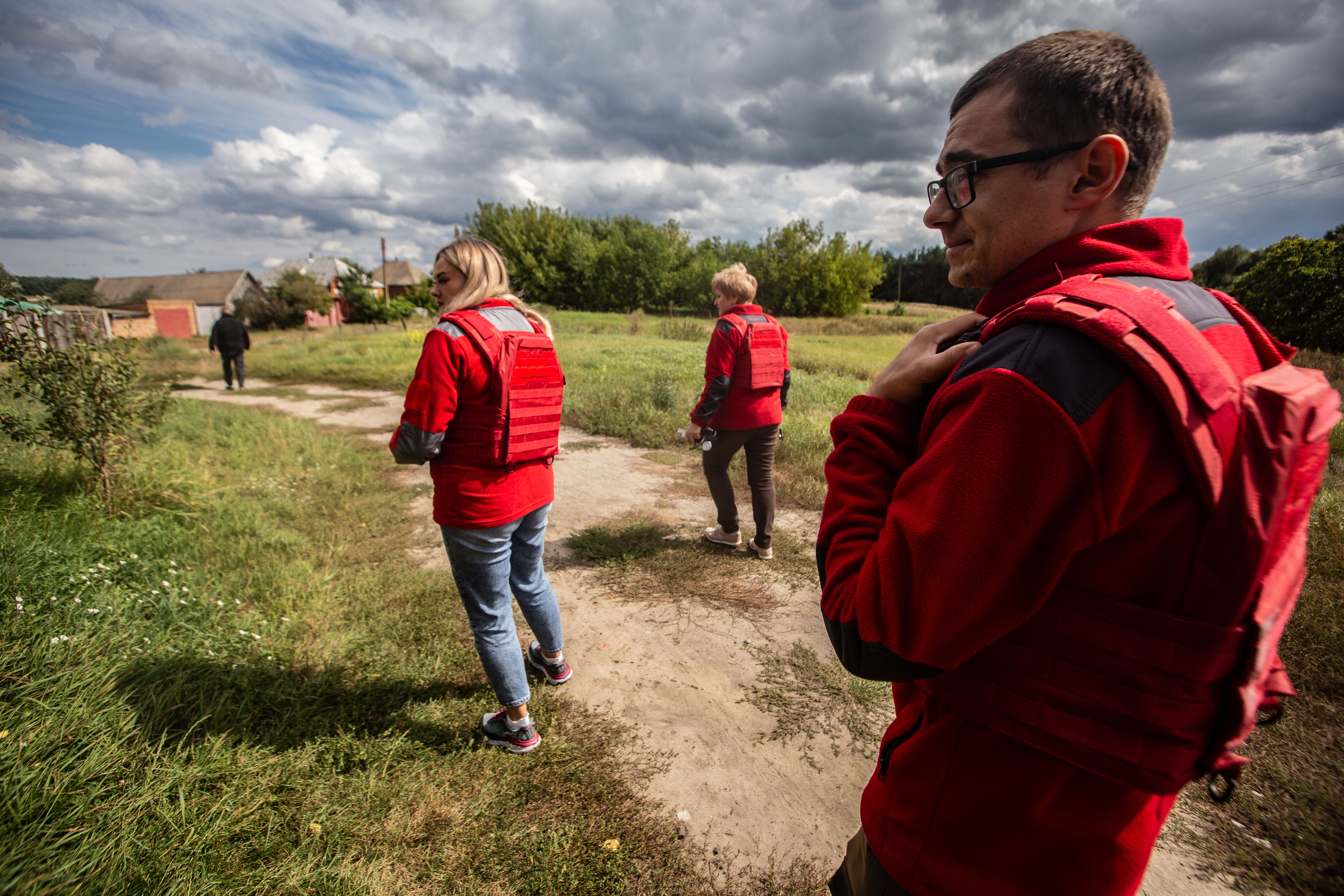 A man and two women walk towards a small village, wearing bright red jumpers over which they have bright red bullet proof vests. They are heading away from the camera but are turned a little towards it so their faces can be partly seen. It is sunny but in the background, there are dark grey rain clouds, giving a moody atmosphere to the photo.