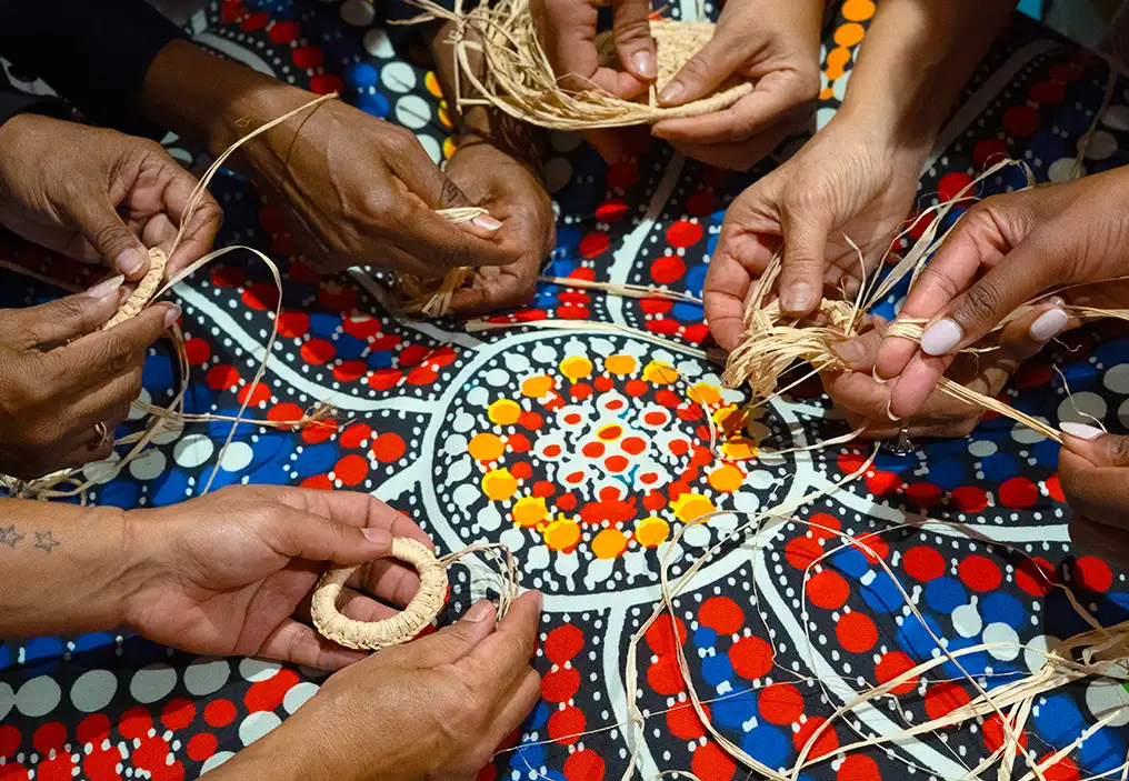 A close up of several pairs of hands weaving natural plant fibres, in a loose circle over a traditional First Nations dot painting with vibrant red, blue, white, yellow and black colouts.