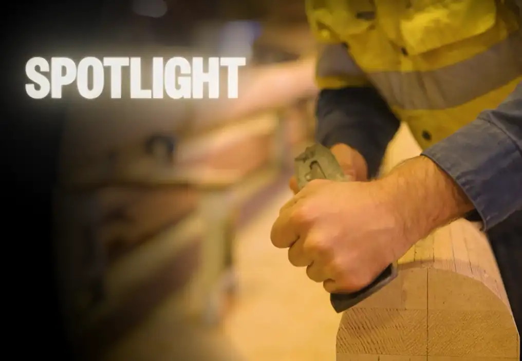A close up of a man’s hands, manually planing a large round mast of light wood. The image is in a circle and surrounded by black, as if illuminated by a spotlight. At the top left is the word ‘SPOTLIGHT’ lightly glowing as though it is a light source.