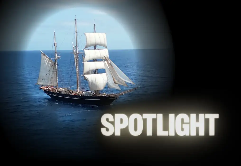 A long shot of the tallship Leeuwin under sail on a calm blue ocean. The photo of the Leeuwin is in a circle and surrounded by black, as if illuminated by a spotlight. At the bottom right is the word ‘SPOTLIGHT’ lightly glowing as though it is a light source.