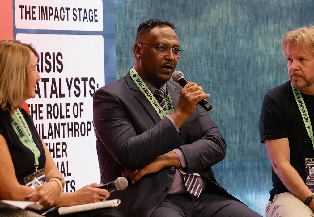A mid shot of Dr Alaeldin Elmalik holding a microphone up to his mouth and in the middle of speaking. He is wearing a charcoal coloured suit and sitting on a tall stool. To the left of frame is Julie Rosenberg, and to the right, Simon Eccleshall. They are both looking at Dr Elmalik.