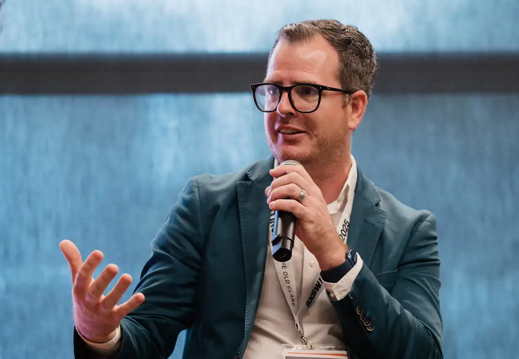 A close up of Ryan Ginard, holding a microphone up with his left hand and gesturing in an open hand, palm up gesture with his right. He is wearing a white shirt and greenish blue jacket, has black rimmed glasses, and has a SXSW Sydney branded lanyard and pass around his neck. Behind him, the blinds shutting out the light from the large windows are a similar blue to his jacket, and are somewhat textured, making a pleasant background.