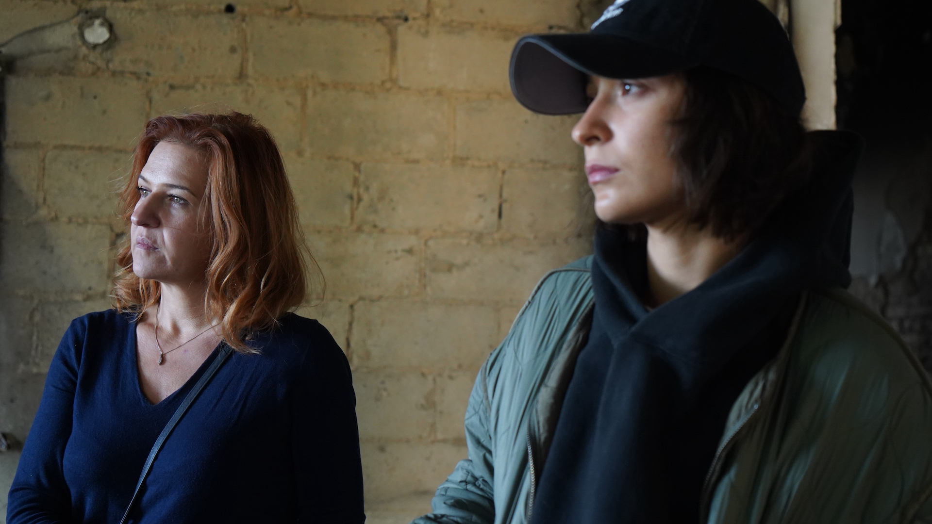 Anya Neistat, with red hair and wearing a navy top, and Solomiia Stasiv, wearing a black baseball cap, a black hoodie and a khaki jacket, look to the left of frame, both looking serious and determined. The background is a wall of rough light-coloured masonry.