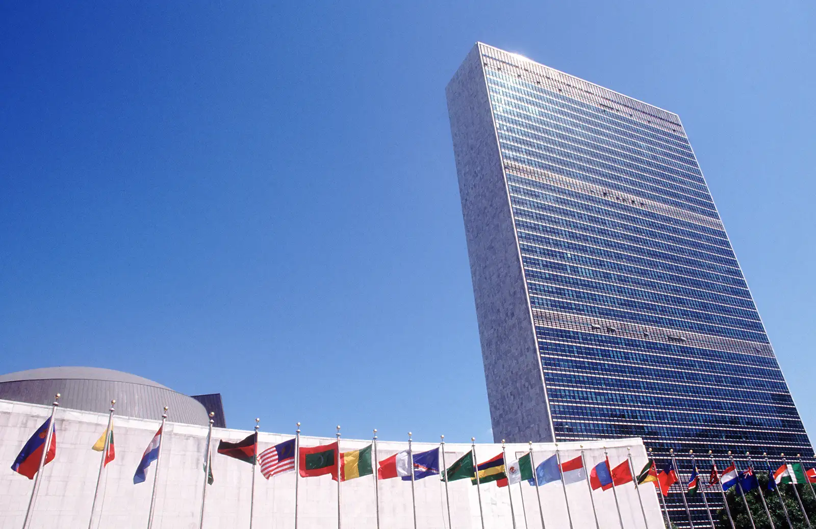 A low angle shot of the UN building in New York city, shot against a bright blue sky with a number of countries flags out the front of it.