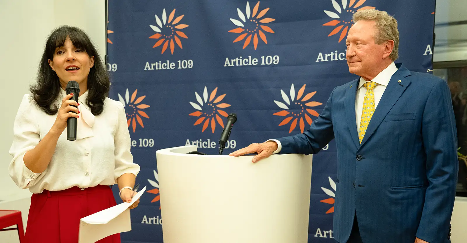 Sofía García, wearing a white blouse and red skirt, speaks into a microphone in front of a blue media wall with the Article 109 logo repeated on it. Also standing in front of the media wall is Andrew Forrest, to the side of a white podium that he is resting his hand on.
