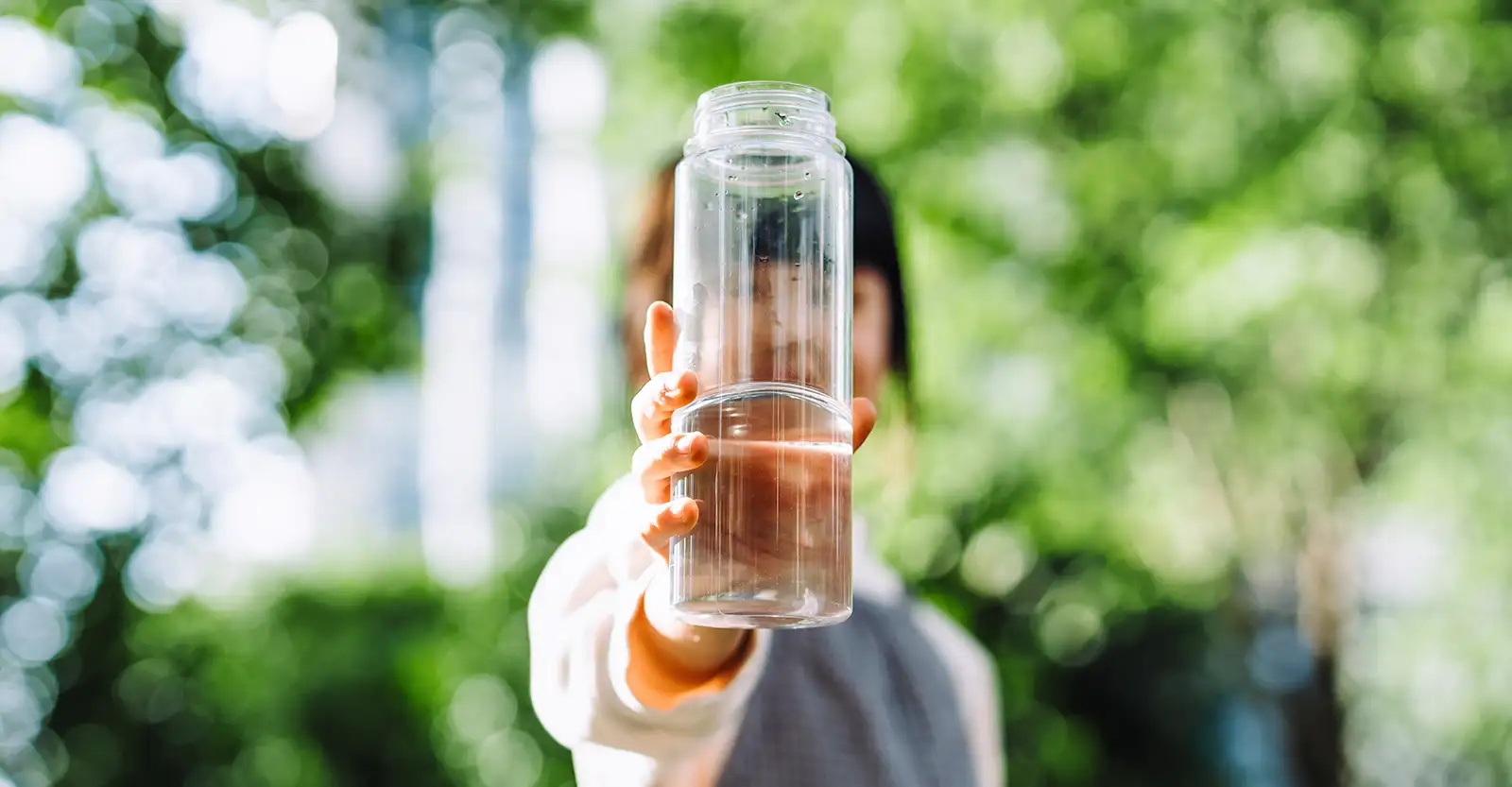 A woman holds a glass bottle out towards the camera. Her face is blurred and obscured by the bottle, so cannot be clearly seen. She is standing in front of a lush green backdrop, but this is also blurred so impossible to make out.
