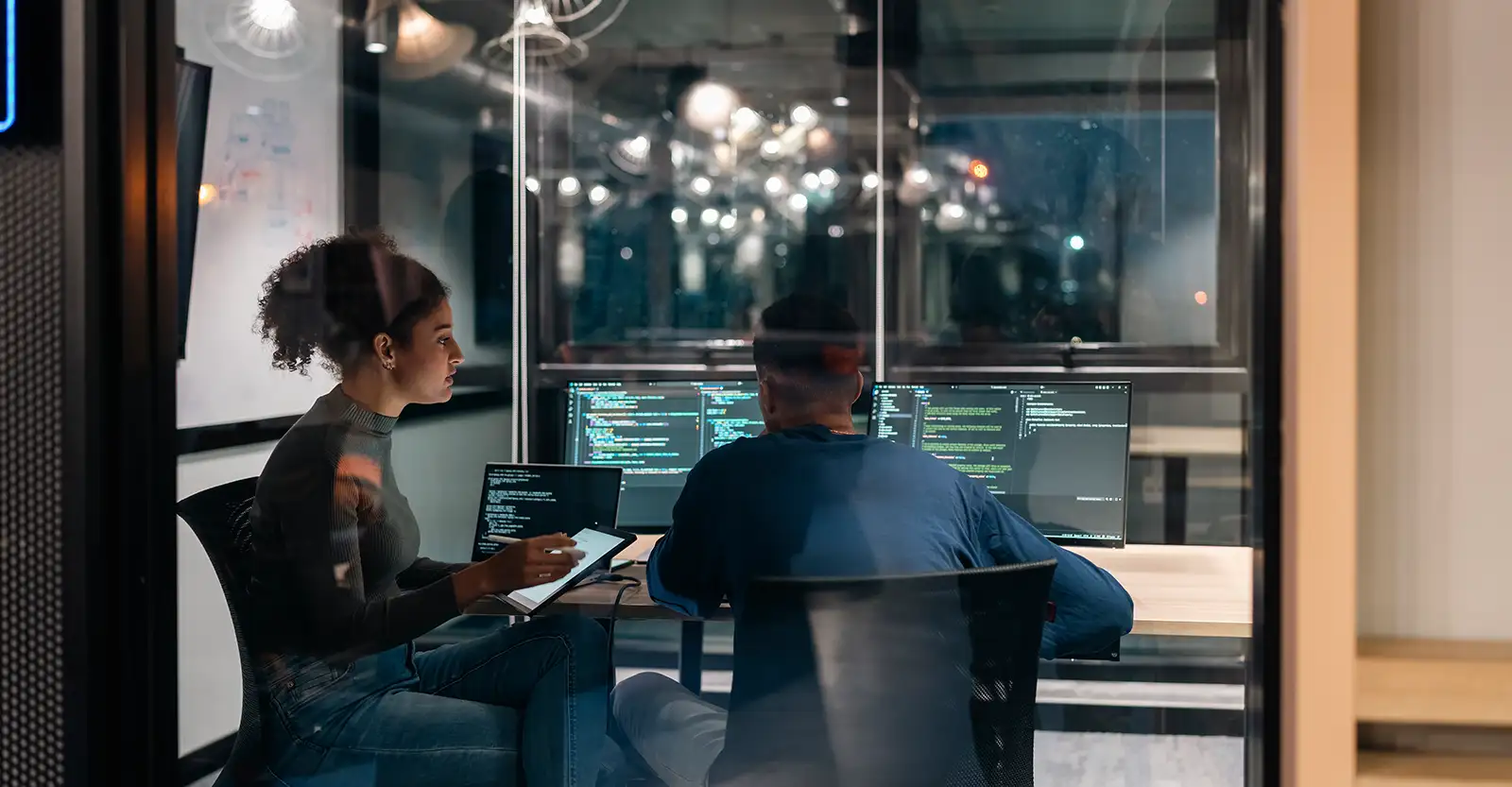 A man and woman sitting in an office wearing casual clothes. The man is sitting with his back to the camera in front of two desktop monitors with code showing on them. The woman, side on to the camera,  has a laptop in front of her showing more code, and is entering something onto a tablet that she is holding.