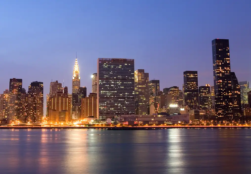 The New York skyline at dusk, with the UN building at the centre of the lit up city skyline. The sky is a dark blue and the river below looks silky from a long exposure and is a darker blue.