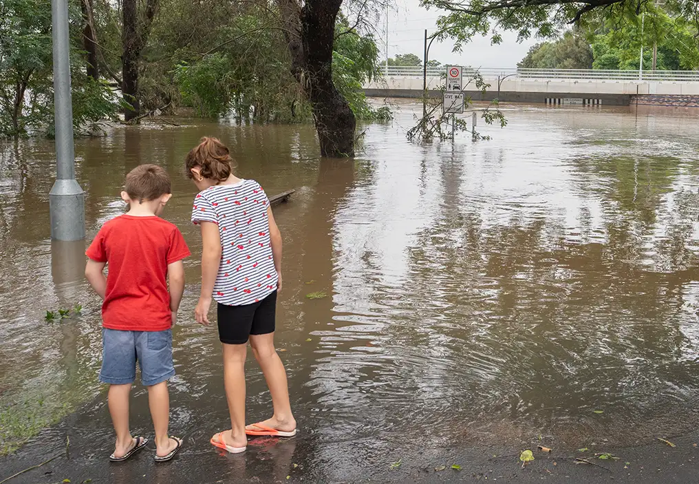A young boy and girl stand with their backs to the camera, looking out over a heavily flooded road, with partially submerged road signs, light poles and trees sticking out of the water. In the background a bridge has escaped being submerged but the water level is past the level of the bottom of the bridge. The children are dressed in shorts, t-shirts and flip-flops and are looking down into the water.