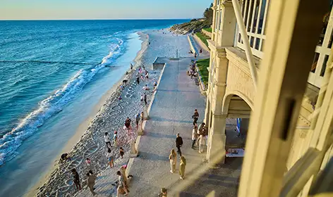 A high angle shot of a beach around sunset. A group of people, children and adults are playing on the narrow strip of sand between the blue water and the sand coloured federation-style building.