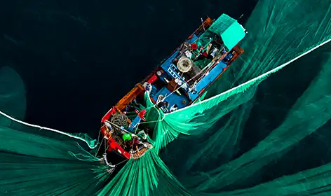 A bird's-eye view of a small fishing trawler with a red hull that has cast out a bright green net. The net is spread out in elegant folds around the boat. The ocean surrounding the boat is dark blue and has no waves.