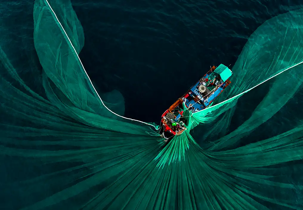 A bird's-eye view of a small fishing trawler with a red hull that has cast out a bright green net. The net is spread out in elegant folds around the boat. The ocean surrounding the boat is dark blue and has no waves.