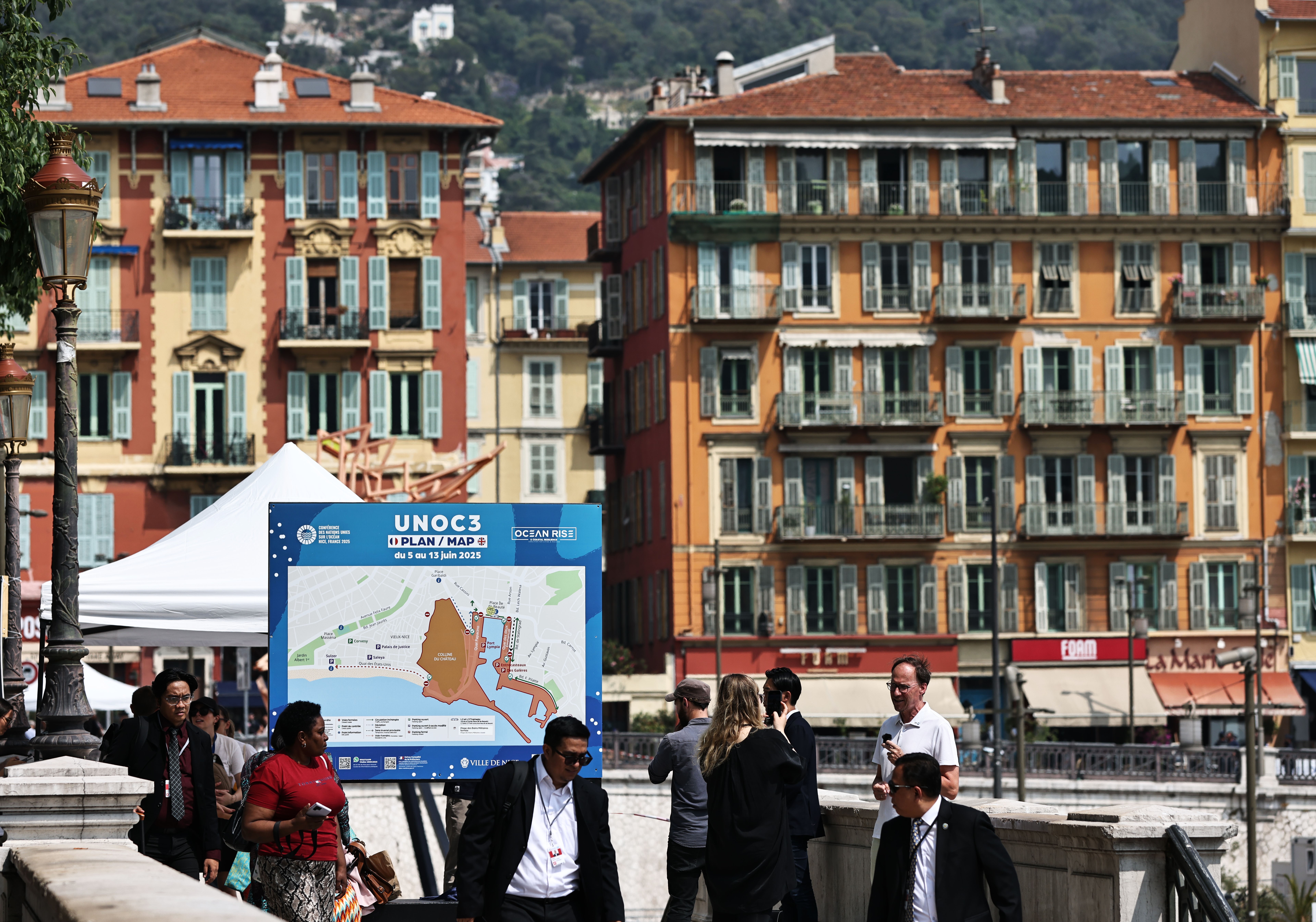 A street with high rise residential buildings in a classic French style in the background. In the foreground are people walking along the street and a large sign that has a map of the UNOC venue.