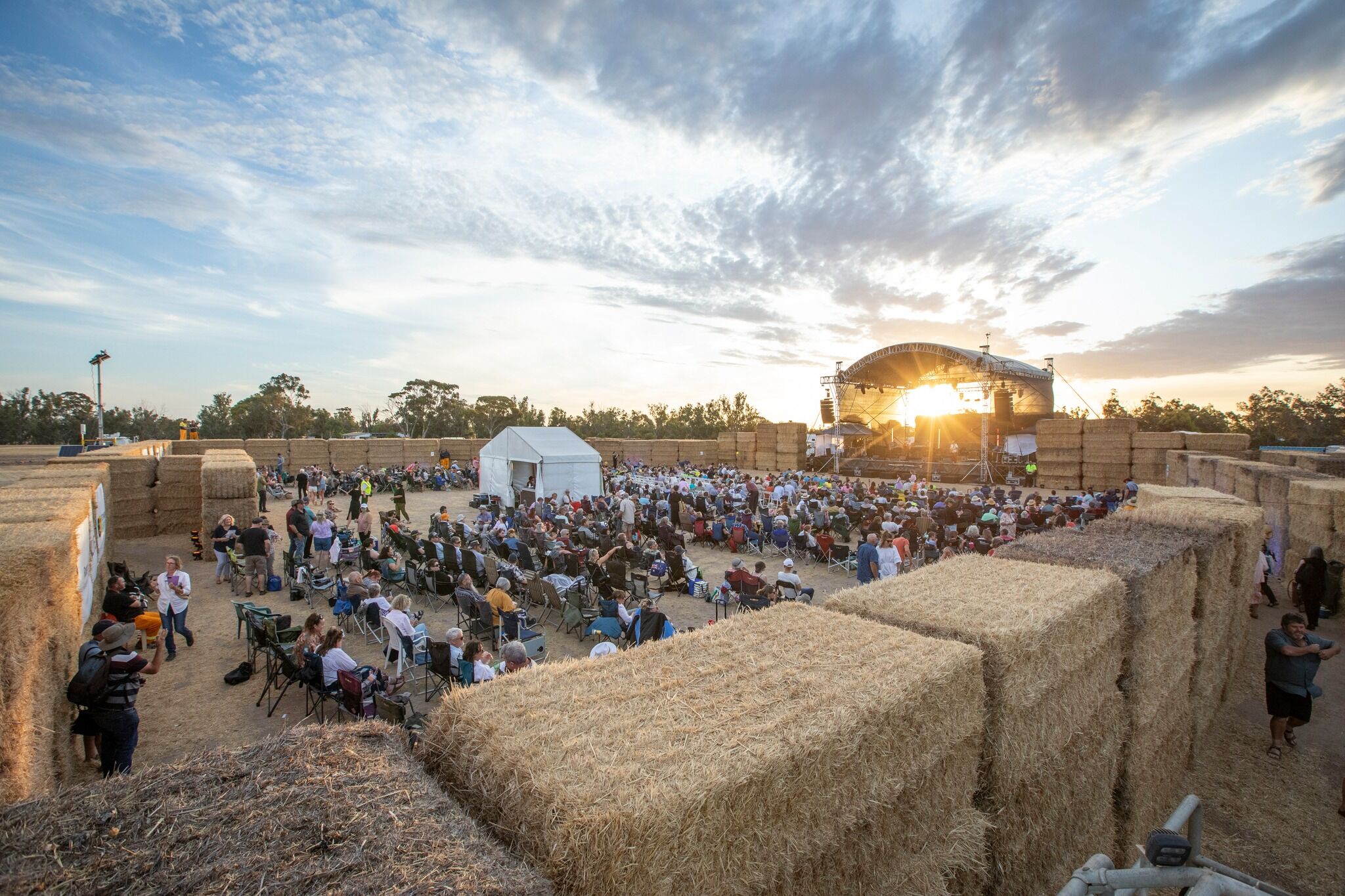 A wide shot of a stage set up in a paddock. The viewing area is bounded by walls of hay bales and inside hundreds of people sitting in camping chairs are waiting for the show to start. The shot has been taken at sunset and the sun is artistically shining through the gap between the stage and the stage roof.