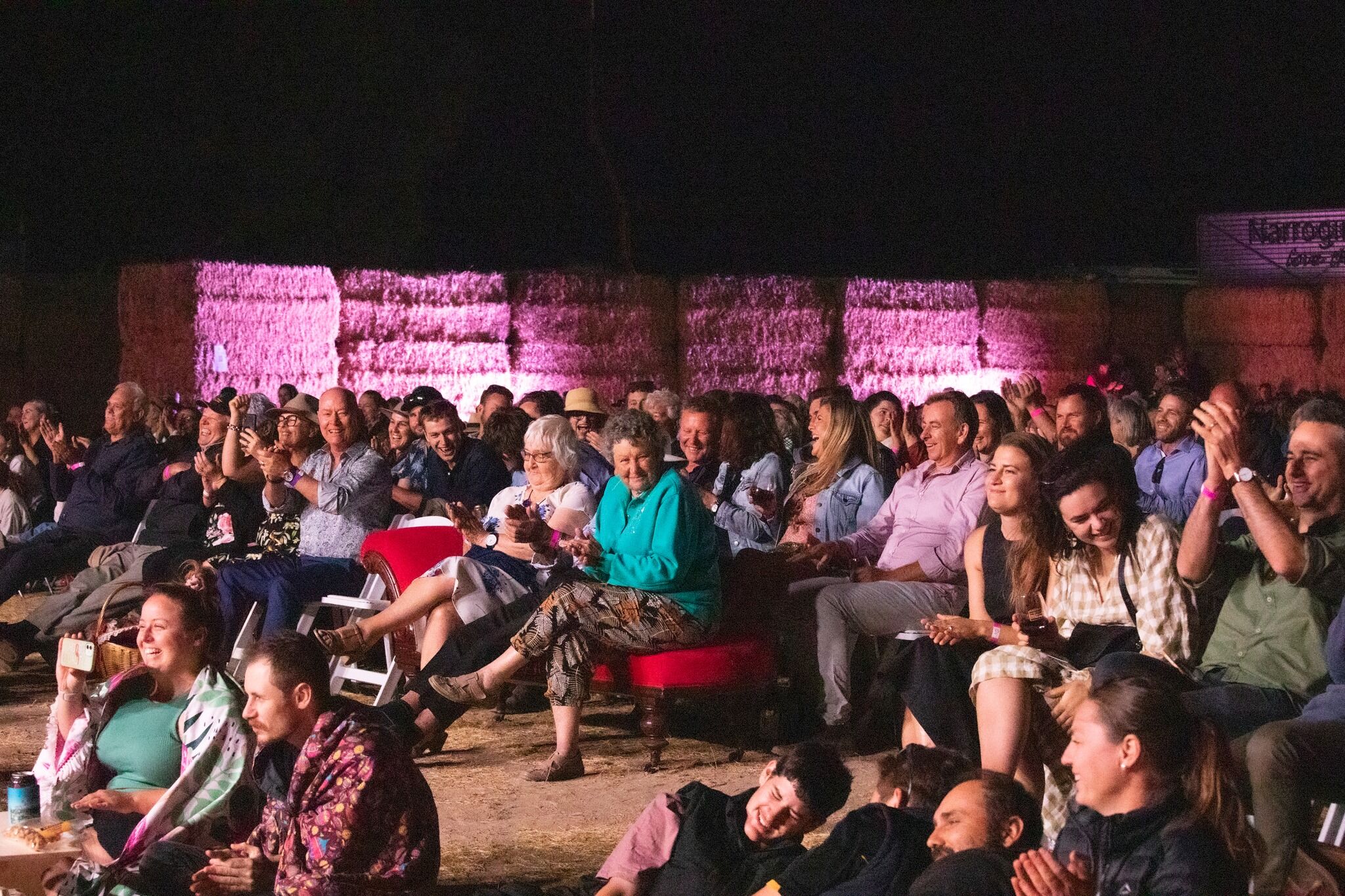 A crowd of people, mostly sitting in camping or outdoor chairs, are smiling and applauding. In the centre of the image, a pair of older women have amusingly brought along a bright red chaise longue instead of a more casual chair. It is night time, and in the background, hay bale walls can be seen, lit up pink from below.