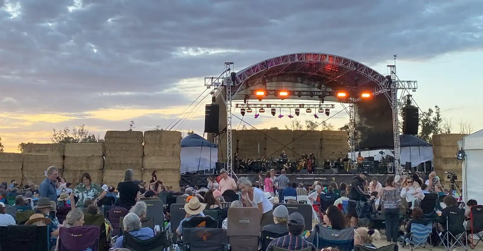 A view of a stage set up outdoors, surrounded by hay bale walls. This photo is taken from the perspective of the audience, some of whom are already sitting and some who are setting up for the outdoor concert.