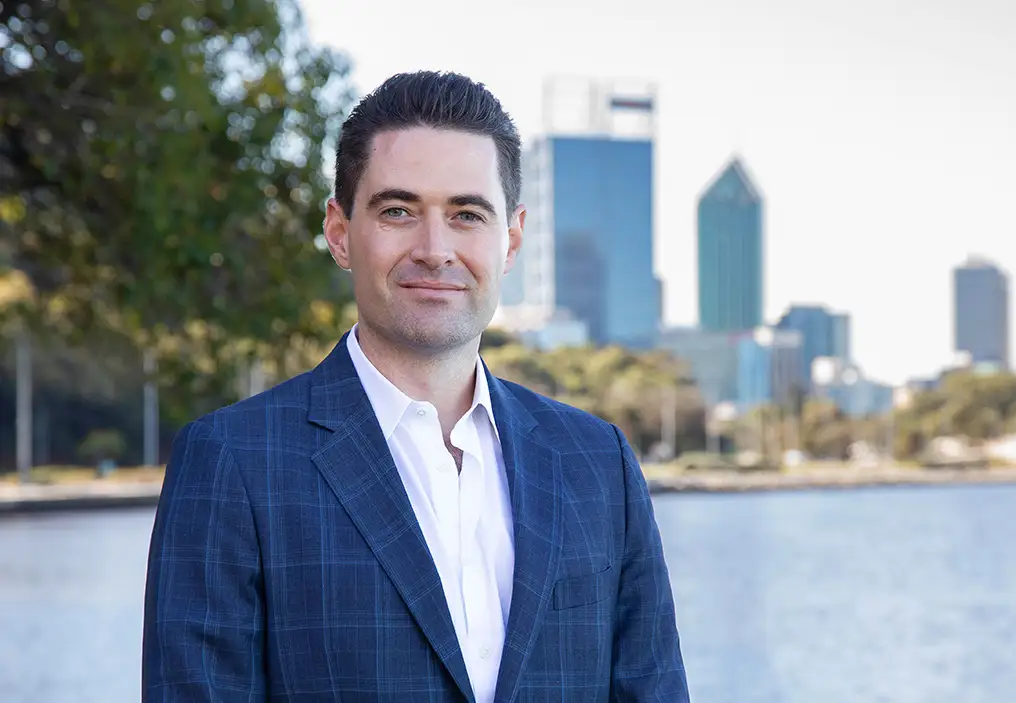 John Hartman wears a mid-blue plaid suit jacket and white shirt, standing in front of the Swan River, with large trees and the city of Perth in soft focus in the background.
