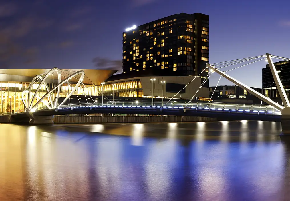 A photo of the Seafarers Bridge and Melbourne Convention Centre taken at night with a slow shutter speed, so the water of the river looks silky and the sky is a beautiful purple. The water reflects purple, blue and gold colours from the sky and the lights.