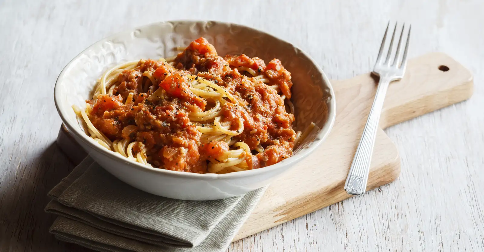 A bowl of spaghetti bolognese in a white rustic patterned ceramic bowl sits on a beige linen napkin on a pine breadboard with shoulders. Next to the bowl is a fork.